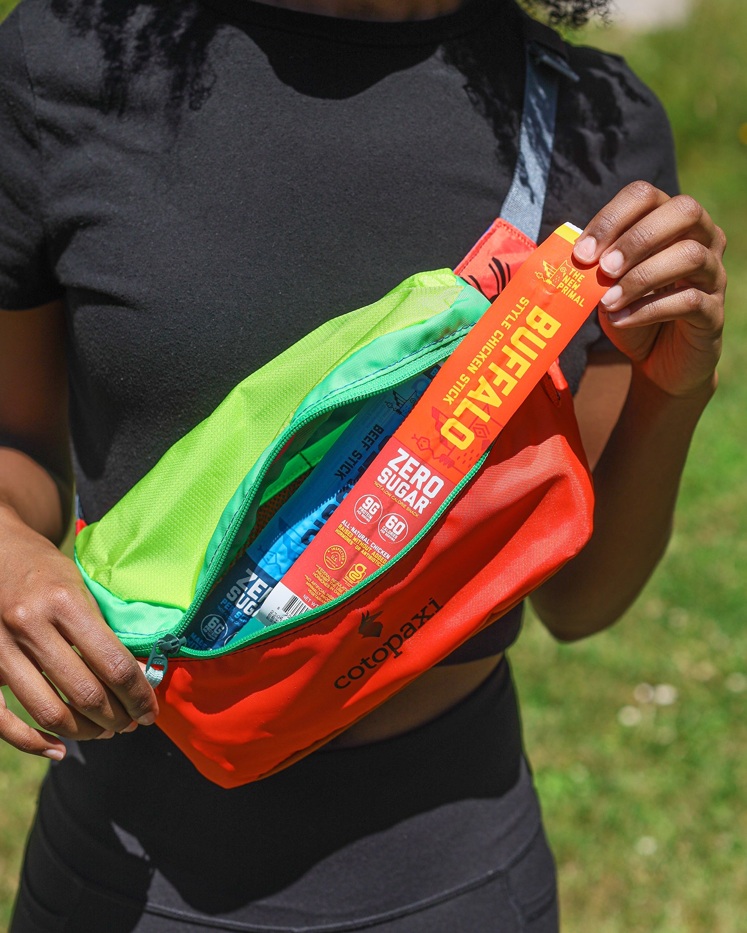 a shot of a woman form the neck down wearing a black tee and black pants holding a multicolored fanny pack taking a buffalo chicken stick out of it.