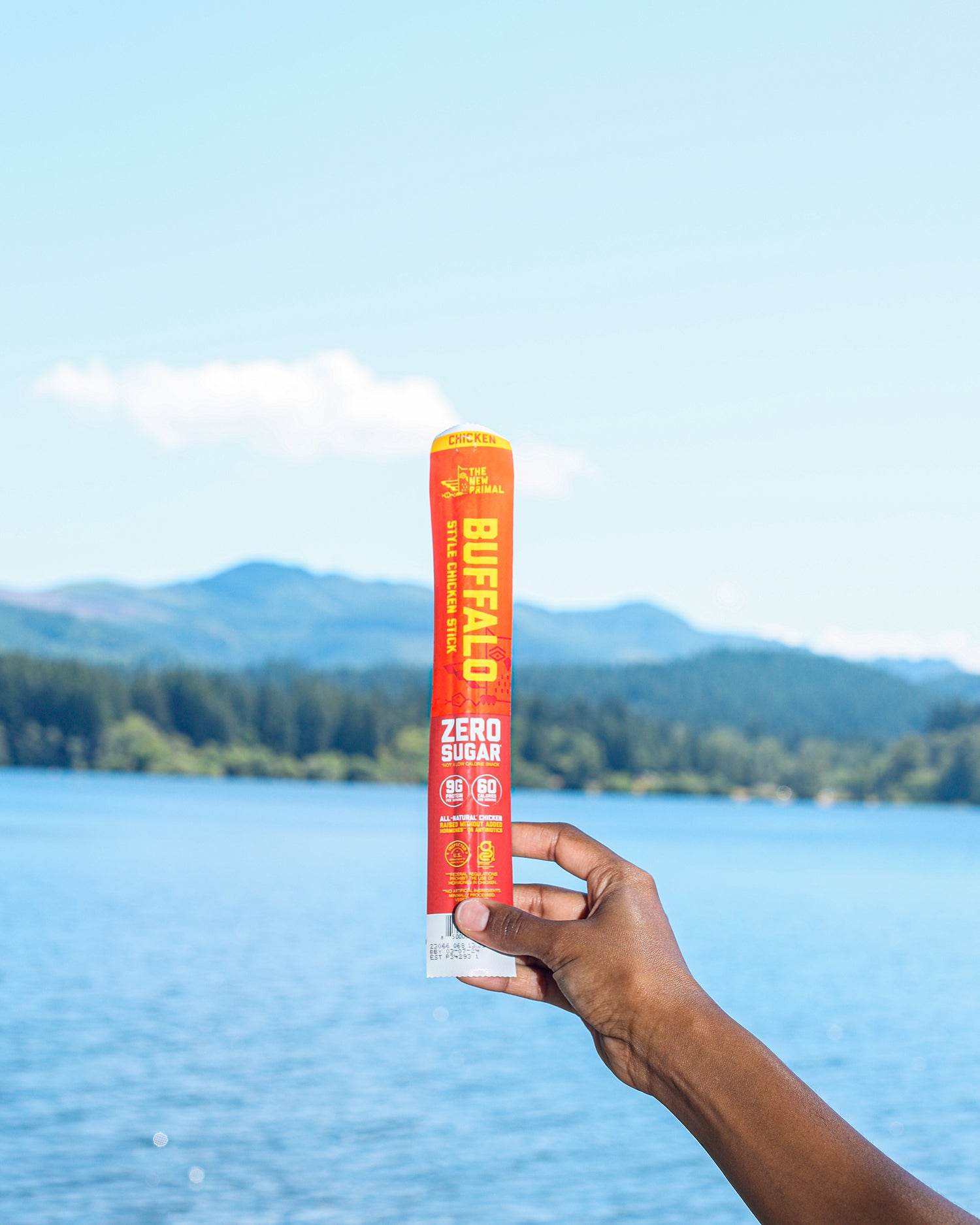 a hand holding an orange and yellow buffalo chicken stick against a lake and mountain view.