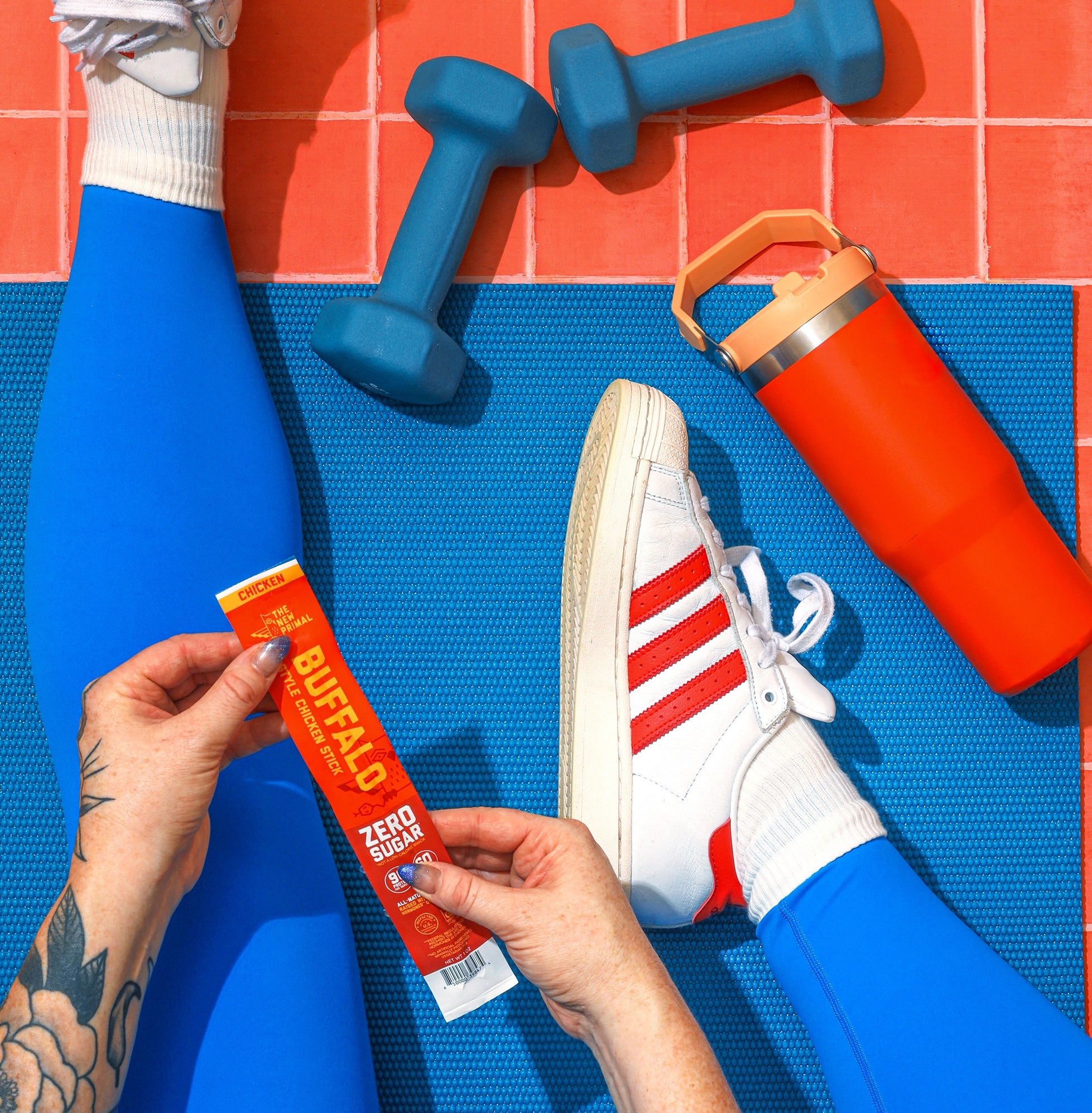  a person in blue leggings and white and red shoes sitting on a blue yoga mat on an orange tile floor with blue weights and an orange water bottle around her holding a buffalo chicken meat stick