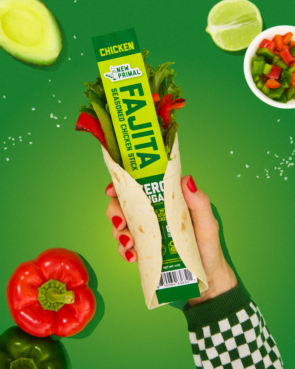 A hand with red nail polish holds a chicken fajita wrap filled with lettuce, red peppers, and tomatoes. The wrap is wrapped in a bright green and white paper sleeve labeled 'New Primal Fajita Seasoned Chicken Stick'.