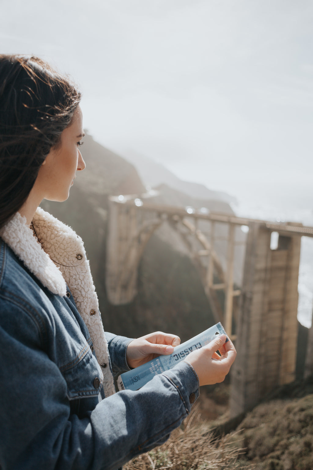 a woman in a denim jacket holding a classic beef stick looking off into the distance in a mountain region