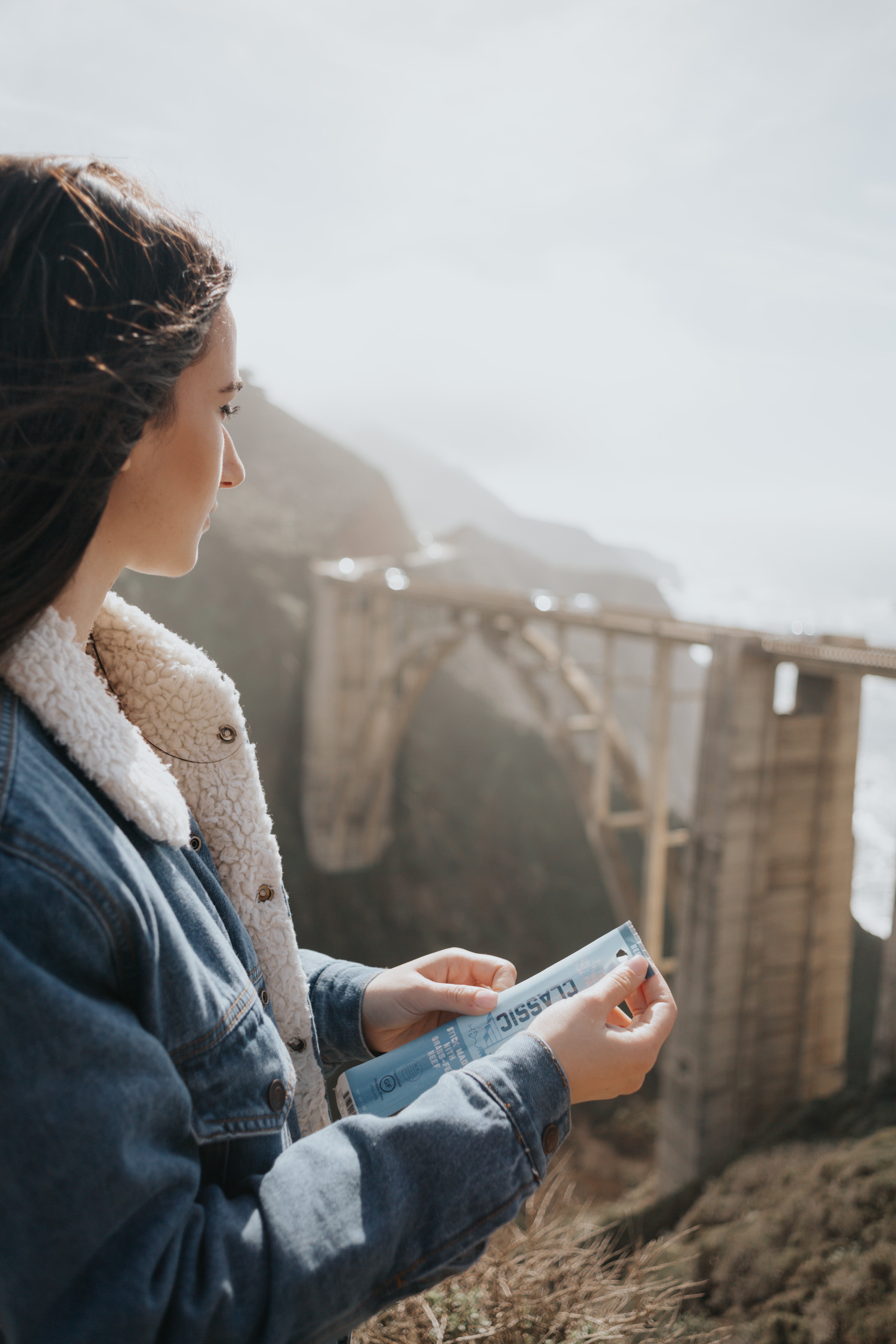 a woman in a denim jacket holding a classic beef stick looking off into the distance in a mountain region